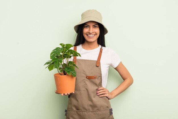 Giovane donna abbastanza ispanica che sorride felicemente con una mano sull'anca e fiducioso concetto di agricoltore o giardiniere