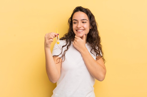 Giovane donna abbastanza ispanica sorridente con un'espressione felice e sicura con la mano sul concetto di termometro a mento