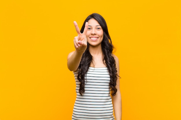 Giovane donna abbastanza latina che sorride e che sembra amichevole, mostrando numero due o secondi con la mano in avanti, conto alla rovescia contro la parete arancione