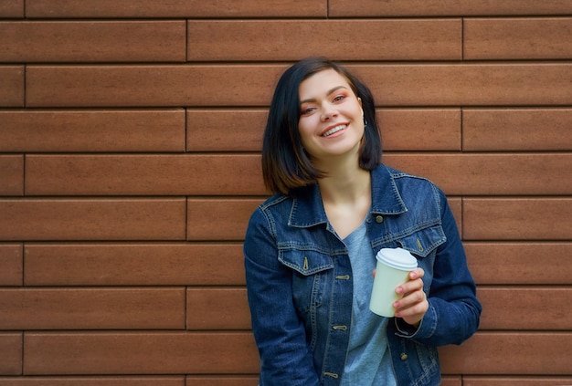 Giovane donna abbastanza mora con tunnel nelle orecchie in giacca di jeans blu in piedi davanti al muro di mattoni godendo nel suo caffè caldo.