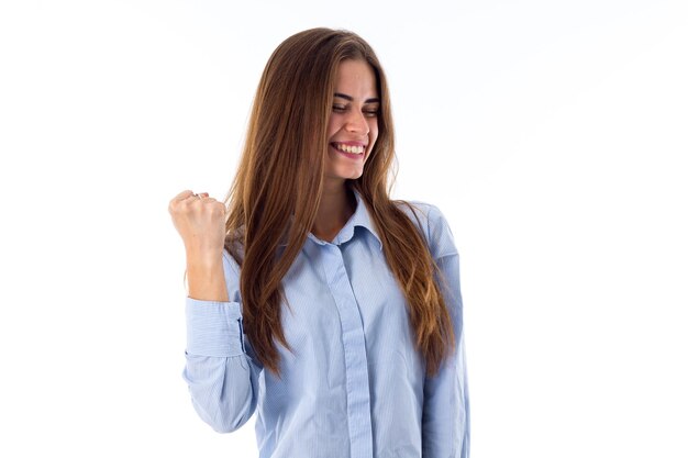 Giovane donna abbastanza sorridente con gli occhi chiusi in camicia blu che mostra il suo pugno su sfondo bianco in studio