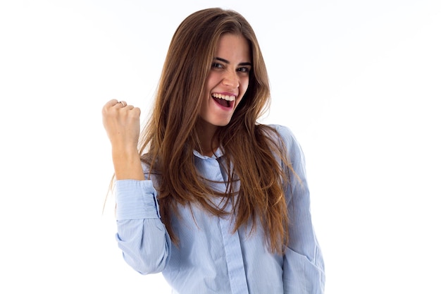 Giovane donna abbastanza sorridente con i capelli lunghi in camicia che mostra il suo pugno su priorità bassa bianca in studio