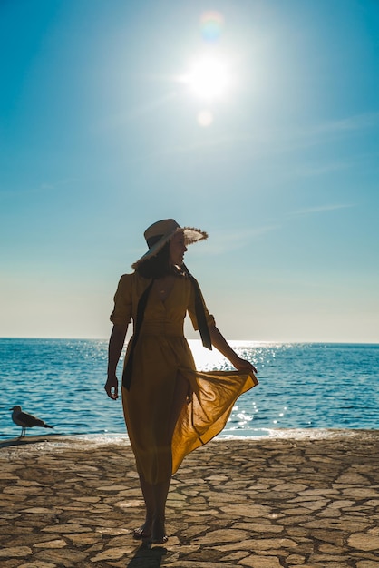 Giovane donna abbastanza sorridente in prendisole giallo che cammina sulla spiaggia di pietra