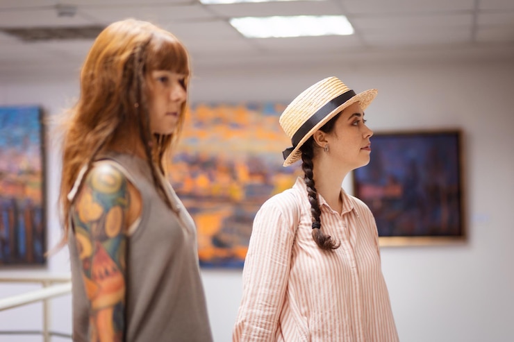 Foto giovane donna abbronzata caucasica con tatuaggio e ragazza che indossa un cappello di paglia che contempla le arti sullo sfondo c'è la sala espositiva della galleria con dipinti concetto di educazione culturale