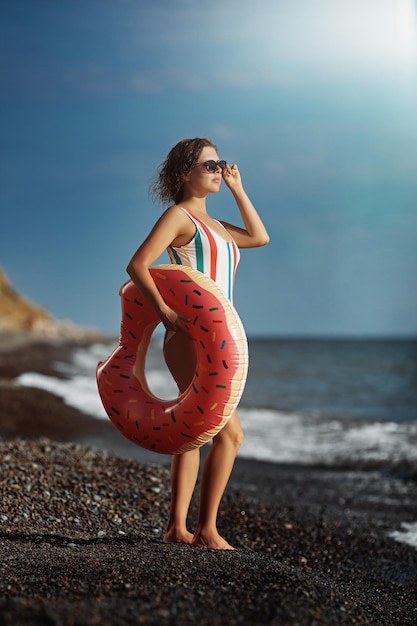 Giovane donna abbronzata in costume da bagno e occhiali da sole in piedi su una spiaggia con una ciambella gonfiabile.