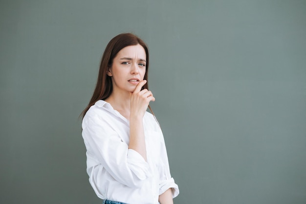 Giovane donna accigliata con i capelli lunghi in camicia bianca su sfondo grigio emozione negativa