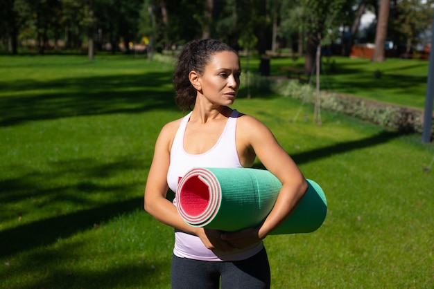 Giovane donna adatta in piedi nel parco e guardando lontano con il tappetino fitness nelle mani