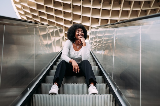 Giovane donna adolescente nera sorridente con i capelli afro che si siede su una scala mobile all'aperto