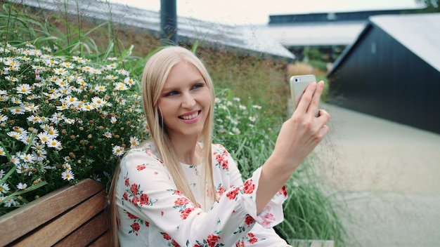 Giovane donna adorabile che per mezzo dello smartphone per fare videochiamata mentre sedendosi sul banco sul tetto vivente del centro commerciale enorme