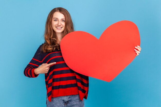 Giovane donna adulta che indossa un maglione in stile casual a righe, in piedi che punta al grande cuore rosso, guardando la fotocamera con un sorriso e un'espressione felice. Studio indoor girato isolato su sfondo blu.