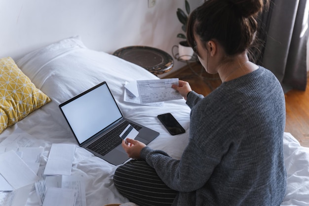 Giovane donna adulta guardando le fatture in possesso di carta davanti al computer portatile con schermo bianco