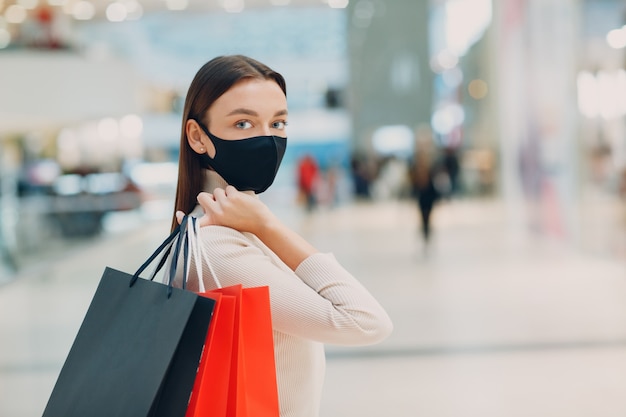 Giovane donna adulta in maschera protettiva medica che trasporta borse della spesa di carta in mano al supermercato