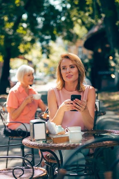 Giovane donna adulta seduta al bar con lo smartphone in mano e distoglie lo sguardo con un sorriso