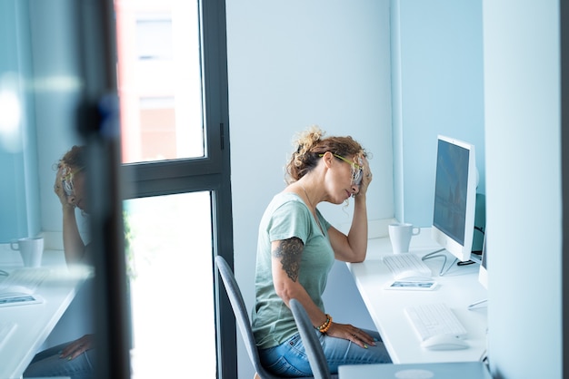 Giovane donna adulta stressata e stanca al desktop davanti al computer e lo stile di vita della giornata di attività lavorativa online toccando la testa per il mal di testa - lavoro d'ufficio - colori della città dell'umore blu