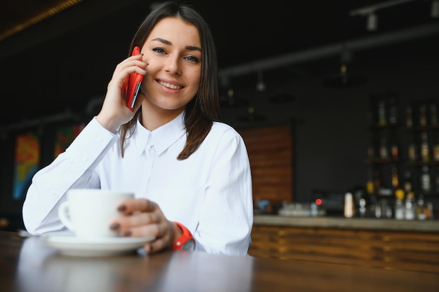 Giovane donna affascinante che chiama con il cellulare mentre è seduta da sola al bar durante il tempo libero, donna attraente con un sorriso carino che parla con il cellulare mentre riposa al bar