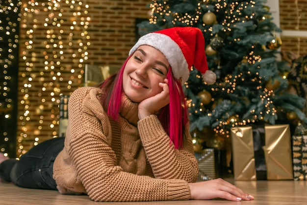 Giovane donna affascinante e sognante con un maglione e un berretto natalizio sul pavimento sullo sfondo di un albero di natale con regali