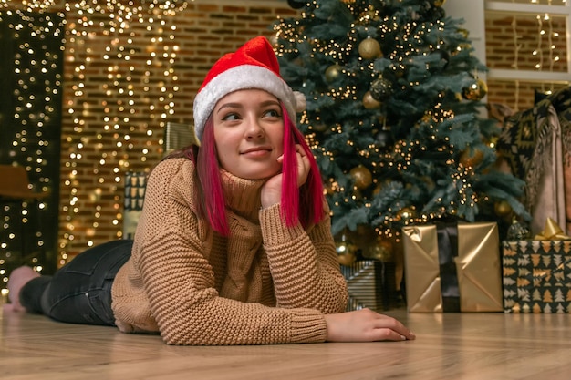 Giovane donna affascinante e sognante con un maglione e un berretto natalizio sul pavimento sullo sfondo di un albero di natale con regali