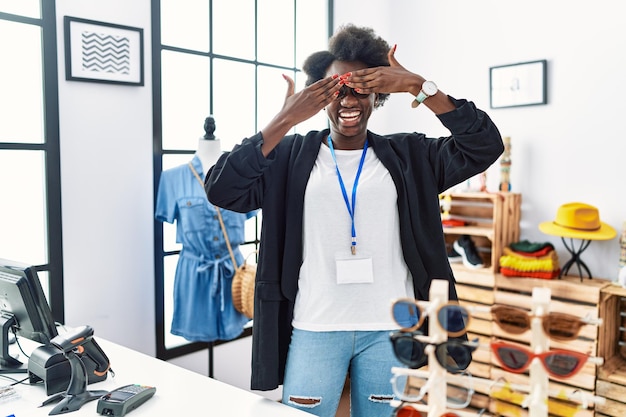 Giovane donna africana che lavora come manager presso la boutique al dettaglio che copre gli occhi con le mani sorridendo allegro e divertente concetto cieco