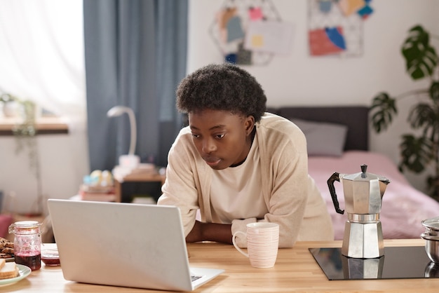 Giovane donna africana che lavora online sul laptop mentre è seduta al tavolo e fa colazione a casa