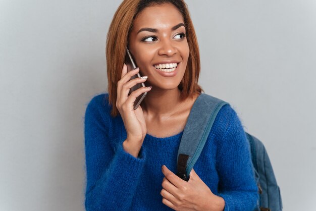 Giovane donna africana felice in maglione con zaino parlando al telefono e guardando da parte.