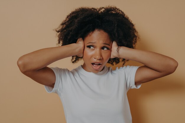 Giovane donna africana infastidita che si copre le orecchie e tiene la bocca aperta isolata su fondo beige