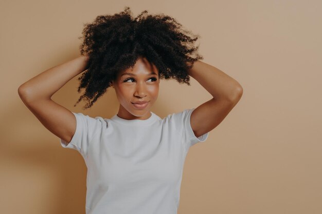 Giovane donna africana sensuale attraente che tocca con i capelli ricci delle mani isolati su fondo beige