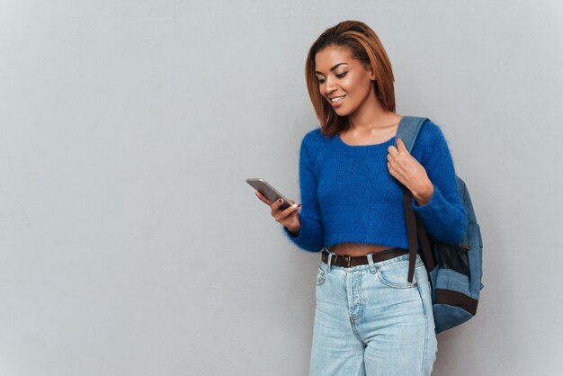 Giovane donna africana sorridente in maglione e jeans con messaggio di scrittura zaino sul telefono.