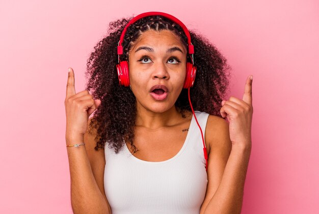 Giovane donna afro-americana che ascolta la musica con le cuffie isolate su sfondo rosa rivolto verso l'alto con la bocca aperta.