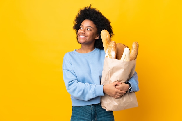 Giovane donna afro-americana che compra qualcosa di pane isolato su giallo guardando mentre sorride