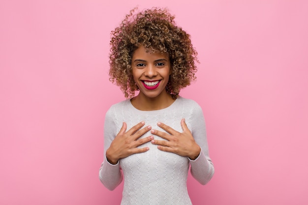 Giovane donna afro-americana che sembra felice, sorpresa, orgogliosa ed eccitata, che punta a sé sul muro rosa