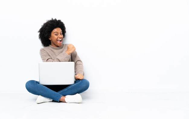 Giovane donna afro-americana con un computer portatile seduto sul pavimento per celebrare una vittoria