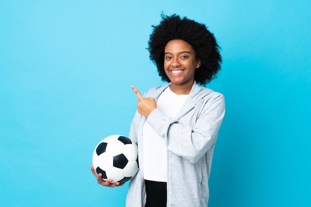 Giovane donna afro-americana isolata su sfondo blu con pallone da calcio e che punta al laterale