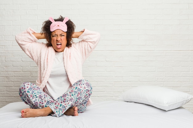 Giovane donna afro-americana nel letto indossando pijama che coprono le orecchie con le mani cercando di non sentire un suono troppo forte.