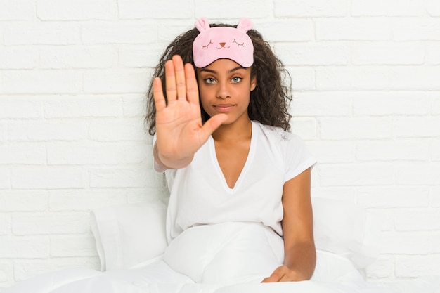 Giovane donna afro-americana seduta sul letto indossando una maschera di sonno in piedi con la mano tesa mostrando il segnale di stop, impedendoti.