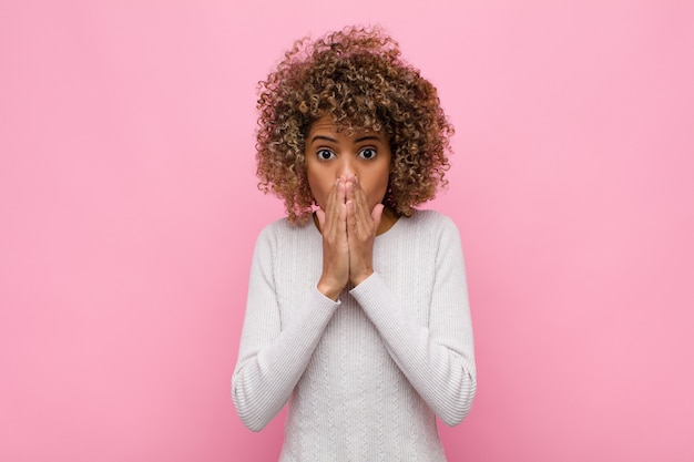 Giovane donna afro-americana sentirsi preoccupata, sconvolta e spaventata, coprendosi la bocca con le mani, guardando ansiosa e avendo incasinato contro il muro rosa