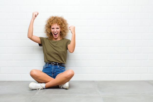 Giovane donna afro che grida trionfalmente, sembrando vincitore eccitato, felice e sorpreso, celebrando seduta su un piano