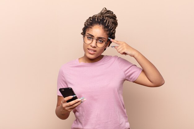 Giovane donna afro che si sente confusa e perplessa, dimostrando che sei pazzo, pazzo o fuori di testa. concetto di smart phone