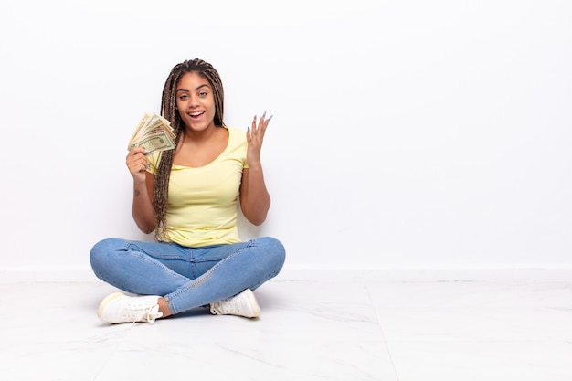 Giovane donna afro che si sente felice, sorpresa e allegra, sorridente con atteggiamento positivo, realizzando una soluzione o un'idea. concetto di denaro