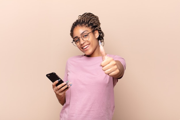 Giovane donna afro che si sente orgogliosa, spensierata, sicura di sé e felice, sorridendo positivamente con il pollice in alto. concetto di smartphone