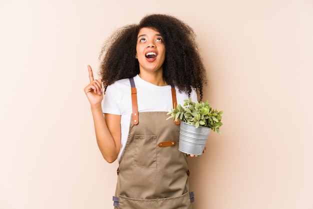 Giovane donna afro giardiniere in possesso di una pianta isolata rivolta verso l'alto con la bocca aperta.