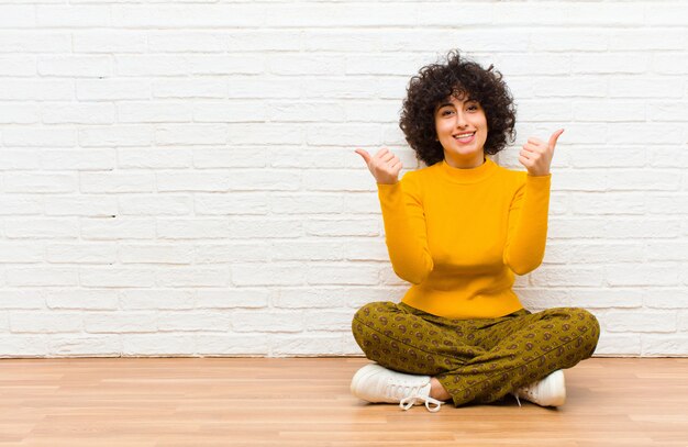 Giovane donna afro graziosa che sorride con gioia e che sembra felice, sentendosi spensierata e positiva con entrambi i pollici in su che si siedono sul pavimento