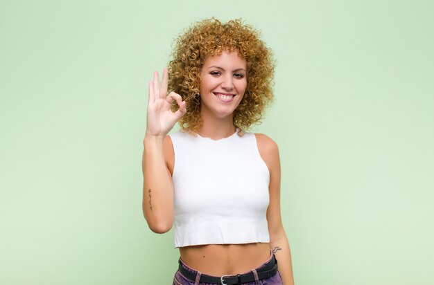 Giovane donna afro sentirsi felice, rilassato e soddisfatto, mostrando l'approvazione con un gesto va bene, sorridendo contro la parete verde