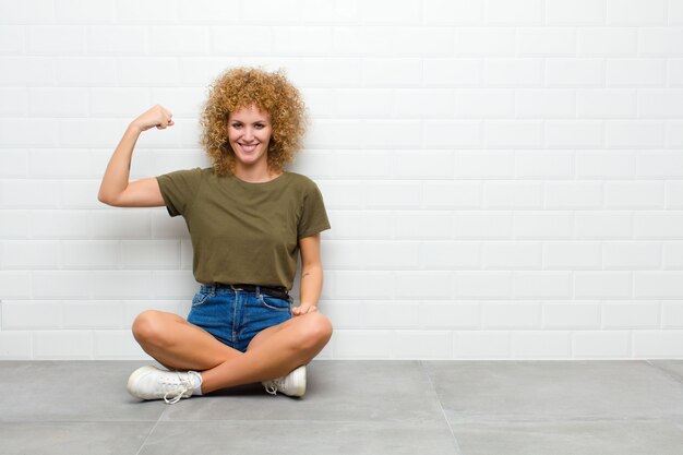 Giovane donna afro sentirsi felice, soddisfatto e potente, flettendo in forma e bicipiti muscolari, guardando forte dopo la palestra seduto su un pavimento