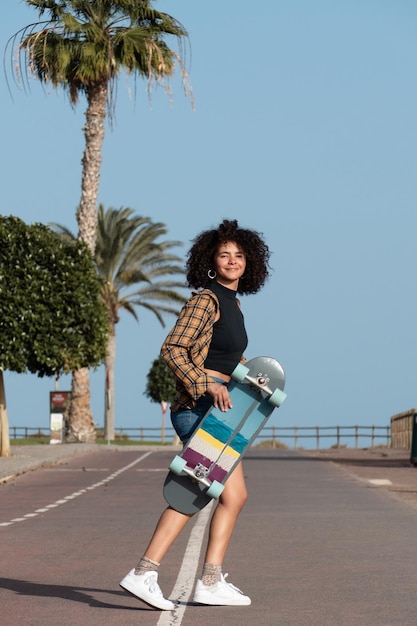 Giovane donna afro skater in piedi all'aperto tenendo il suo skateboard e sorridente