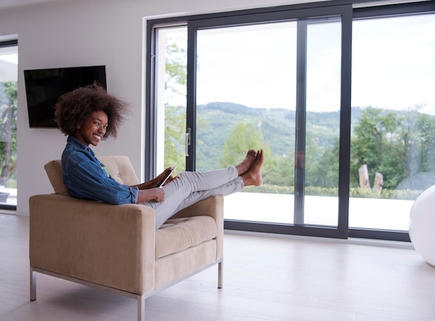 Giovane donna afroamericana a casa rilassante nel suo soggiorno di lusso leggendo un tablet PC digitale navigare in internet e lavorare
