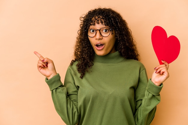 Giovane donna afroamericana afro che tiene un cuore di giorno di biglietti di S. Valentino che indica al lato