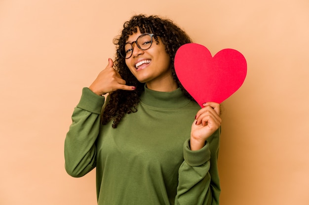 Giovane donna afroamericana afro che tiene un cuore di San Valentino che mostra un gesto di chiamata di cellulare con le dita.