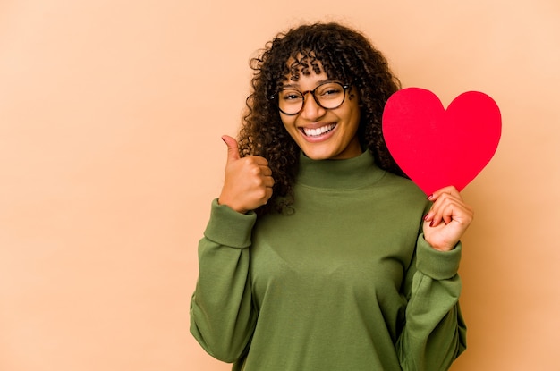 Giovane donna afroamericana afro che tiene un cuore di San Valentino sorridendo e alzando il pollice