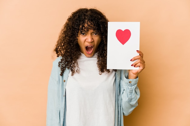 Giovane donna afroamericana afro che tiene una carta di San Valentino urlando molto arrabbiata e aggressiva.