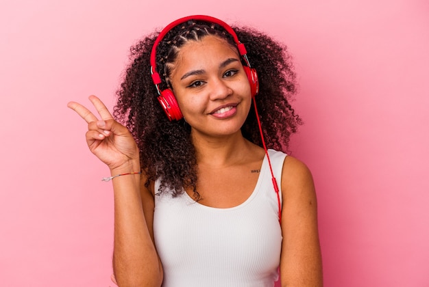Giovane donna afroamericana che ascolta la musica con le cuffie isolate su sfondo rosa gioiosa e spensierata che mostra un simbolo di pace con le dita.